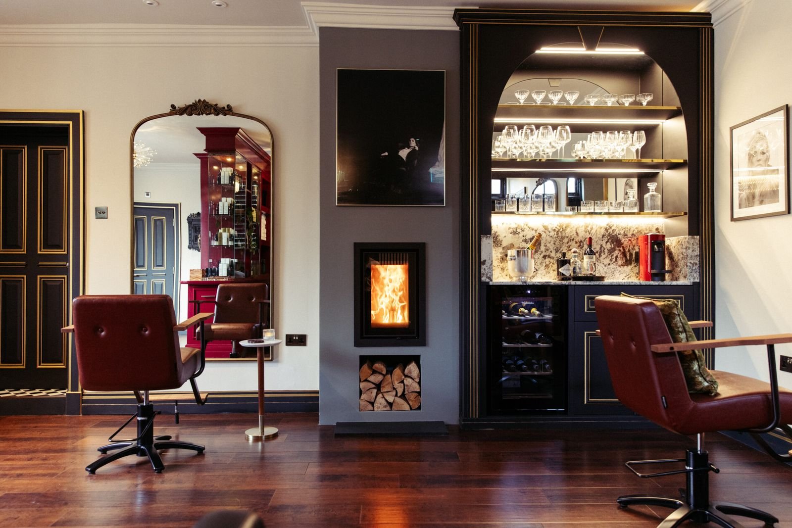Salon interior with two styling chairs, a lit log fire and a glass-fronted drinks cabinet